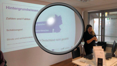 Ramona Armbrust steht vor einer Wand auf der eine Bildschirmpräsentation ist. Auf der Folie steht "Hintergrundwissen, Zahlen und Fakten". In der Mitte des Bildes ist eine große Lupe eingefügt, die einen Teil des Fotos vergrößert.