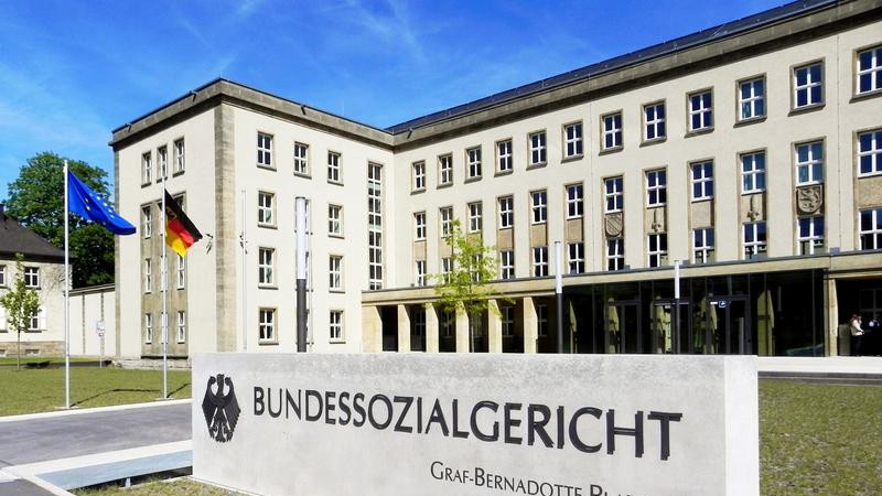 Das Foto zeigt den Eingangsbereich zum Bundessozialgericht. Eingrußes weißes Gebäude. Davor befindet sich eine weiße Tafel mit der Aufschrift Bundessozialgericht.